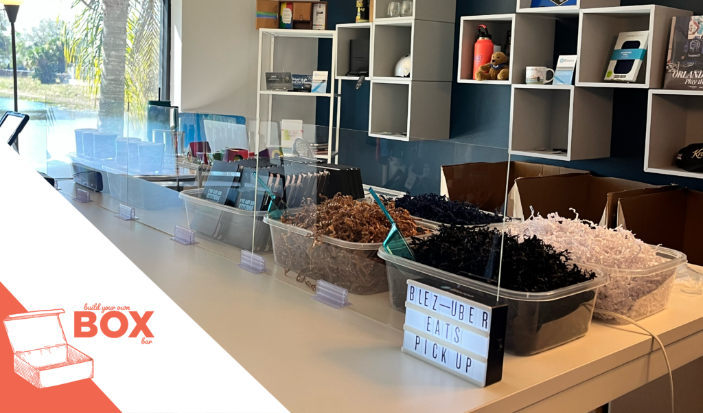 A Blezoo build-your-own-box kitting station at an in-house event, featuring a long table with trays of crinkle paper in multiple colors, display shelves with branded promotional products